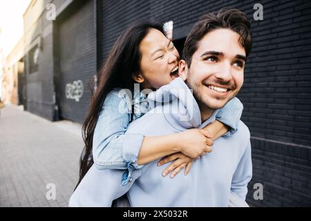 Giovane uomo che porta la sua ragazza sulla schiena mentre cerca di mordergli l'orecchio, camminando per la strada della città. Un paio di salvadanaio in città. Foto Stock