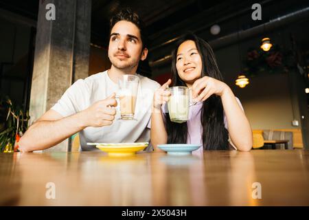 Una coppia seduta in un bar che guarda lontano e sorride. Coppia in un appuntamento in un bar locale che beve caffè e tè matcha. Foto Stock