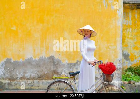 Donna vietnamita in una tradizionale Ao dai con la sua bicicletta in piedi davanti a un muro giallo, Hanoi, Vietnam Foto Stock