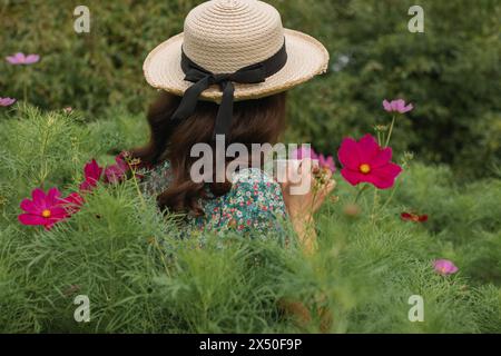 Vista posteriore di una giovane donna che indossa un cappello di paglia e un abito estivo, in piedi in un prato estivo bevendo una tazza di tè, Bielorussia Foto Stock