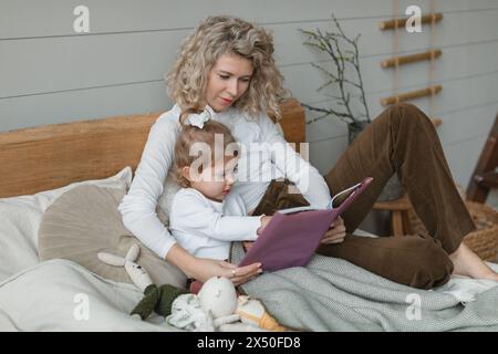 Madre e figlia sedute a letto a leggere un libro Foto Stock