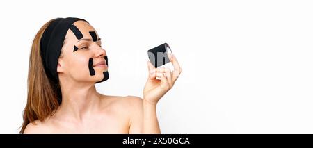 La modella di bellezza con nastri facciali kinesio sul viso guarda un rotolo di nastro k in mano e sorride davanti allo sfondo bianco con spazio di copia. Foto Stock