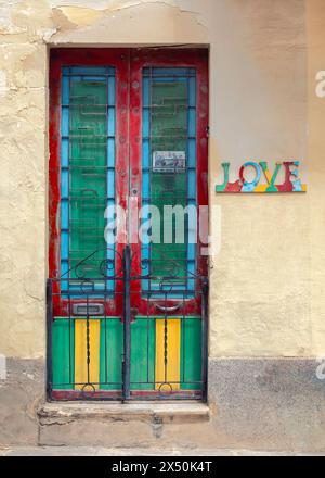 Una porta maltese vibrante e intemprata, adornata da pannelli colorati contro una rustica parete gialla. Sul muro accanto c'è un cartello che dice "amore". Foto Stock