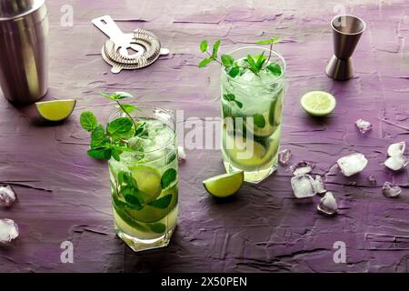 Cocktail Mojito, due bicchieri. Bevanda fredda estiva con lime, menta fresca e ghiaccio. Raffreddare la bevanda su uno sfondo viola con accessori per la miscelazione Foto Stock