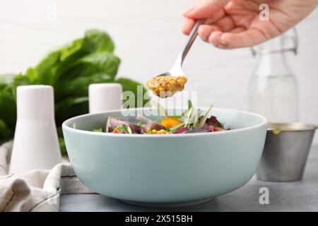 Donna che versa una gustosa salsa a base di aceto (Vinaigrette) dal cucchiaio alla ciotola con insalata al tavolo grigio, primo piano Foto Stock