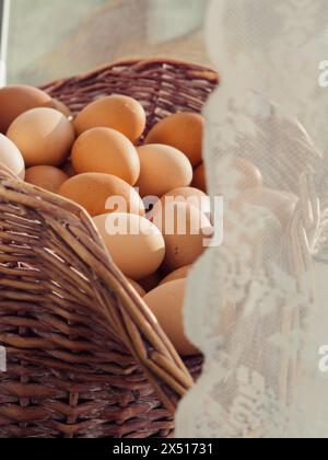 Primo piano verticale di un grande cesto pieno di uova su un davanzale dietro tende bianche con lacci. Foto Stock