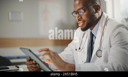 Felice e sorridente medico afroamericano maschio che indossa il cappotto bianco che lavora su un tablet computer nel suo ufficio. Operatore sanitario medico che lavora con i risultati dei test, pianificazione del trattamento del paziente. Foto Stock
