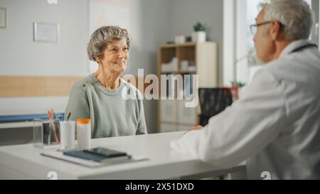 Il medico di famiglia di mezza età sta parlando con la paziente anziana durante la consultazione in una clinica sanitaria. Medico caucasico esperto in camice da laboratorio seduto dietro una scrivania di computer in ufficio ospedaliero. Foto Stock