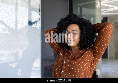 Una giovane donna afroamericana gode di un momento di pace in un ufficio luminoso e moderno. Si appoggia di nuovo sulla sedia con gli occhi chiusi, mostrando un senso di rilassamento e soddisfazione. Foto Stock