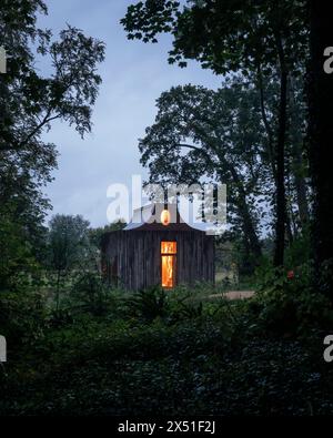 Il padiglione dell'Apiario visto al crepuscolo, con alberi circostanti che si stagliano contro il cielo. le luci interne sono dorate e mostrano la finestra dell'atrio. Il Beezantiu Foto Stock