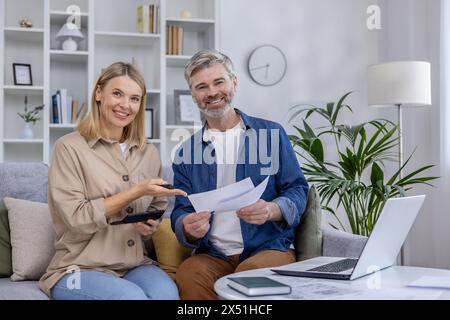 Una coppia matura seduta su un divano, sorridendo mentre discutono documenti e piani finanziari insieme, con una calcolatrice e un portatile in un moderno salotto. Foto Stock