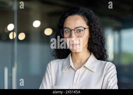 Una donna d'affari ispanica che indossa gli occhiali si trova in un ambiente d'ufficio, trasudando professionalità e determinazione. Foto Stock