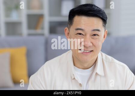 Ritratto di un uomo asiatico felice di mezza età seduto in un salotto contemporaneo, che indossa una camicia casual color crema con un sorriso rilassato e sicuro. Foto Stock