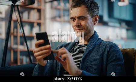 Uomo caucasico eccitato che usa lo smartphone mentre riposa su un divano nell'elegante soggiorno. Happy Man sorride a casa e chiacchiera con colleghi e clienti tramite Internet. Utilizzo dei social network. Foto Stock