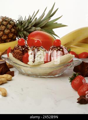 Triplo sundae di banana split con salsa fudge calda, arachidi, fragole, ciliegie su superficie di marmo, circondati da ingredienti. Foto Stock