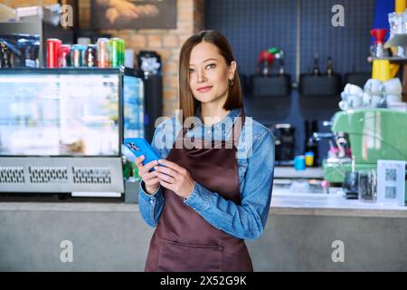 Giovane lavoratrice di servizio in grembiule con smartphone nel ristorante e nella caffetteria Foto Stock
