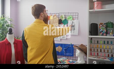 Un uomo barbuto con una camicia gialla indica uno schizzo di moda in un negozio di sartoria ben organizzato con bobine di filo colorato. Foto Stock