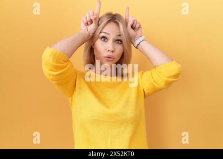 Giovane donna caucasica che indossa un maglione giallo che fa un gesto divertente con il dito sopra la testa come corna di toro Foto Stock