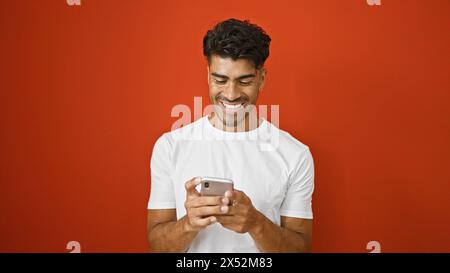 Bel giovane ispanico in camicia bianca sorridente al telefono su sfondo rosso isolato Foto Stock