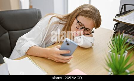 Una giovane donna si appoggia su una scrivania da ufficio, utilizzando il suo smartphone mentre è circondata da accessori di lavoro. Foto Stock