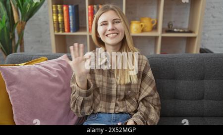 Una giovane donna bionda sorridente ondeggia alla telecamera in un accogliente soggiorno con arredamento moderno Foto Stock