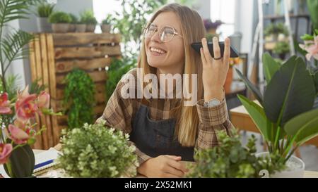 Giovane donna bionda sorridente che ascolta un messaggio vocale all'interno di un negozio di fiori verde, che indica un contesto floreale aziendale. Foto Stock