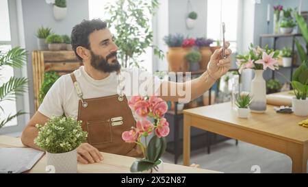 Uomo barbuto che scatta un selfie in un vivace negozio di fiori pieno di piante verdi e fiori colorati. Foto Stock