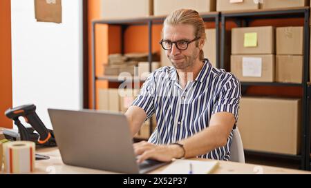 Un uomo sicuro di sé con gli occhiali lavora su un computer portatile in un moderno ufficio di magazzino, circondato da scaffali e scatole. Foto Stock
