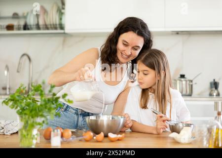 La mamma aggiunge il latte al recipiente, la ragazza mescola gli ingredienti con la frusta. Foto Stock