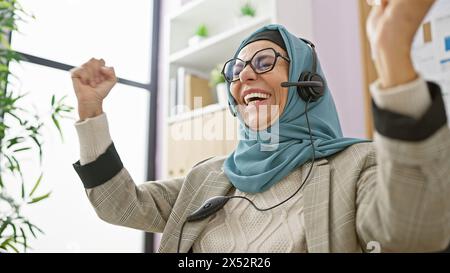 Una donna di mezza età che indossa un hijab festeggia in ufficio con le cuffie indossate. Foto Stock