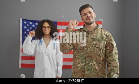 Una donna medico e un uomo soldato con pugni alzati davanti a una bandiera americana al chiuso Foto Stock