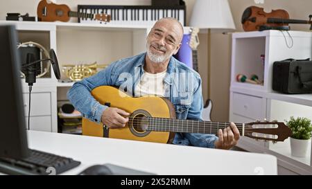 Un uomo maturo sorridente suona la chitarra in un moderno studio domestico pieno di strumenti musicali e apparecchiature di registrazione. Foto Stock