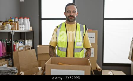 Un giovane ispanico con la barba che indossa un giubbotto "volontario" si trova in un magazzino coperto pieno di scatole. Foto Stock