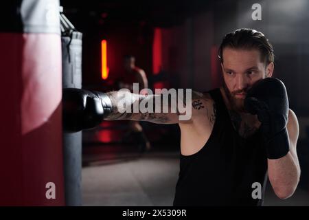 Uomo muscoloso con tatuaggi su braccio e petto in piedi davanti al sacchetto di pugni e colpirlo durante l'allenamento in palestra o nell'anello di pugilato Foto Stock