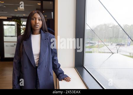 Questa immagine cattura una giovane donna afroamericana in un ambiente professionale, vestita con un abito da lavoro a righe, in piedi accanto a una grande finestra con vista di una giornata di pioggia all'aperto. Il suo comportamento calmo e il suo aspetto elegante trasmettono fiducia e professionalità in un ambiente aziendale moderno. Giovane donna d'affari afro-americana alla finestra nel moderno ufficio durante il giorno della pioggia. Foto di alta qualità Foto Stock