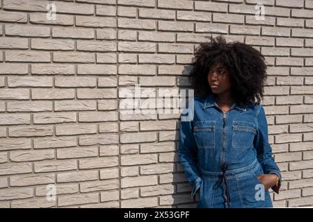 Questa foto candida mostra una giovane donna nera con un'acconciatura afro, vestita con una tuta in denim alla moda. Si appoggia su un muro di mattoni bianchi, la sua posizione trasuda un atteggiamento rilassato ma sicuro di sé, evocando la moda urbana e l'individualità. Elegante donna con outfit in denim appoggiata al muro in mattoni. Foto di alta qualità Foto Stock