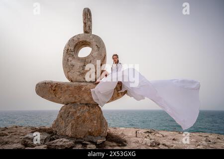 Una donna siede su una scultura in pietra fatta di grandi pietre. È vestita con un lungo vestito bianco, sullo sfondo del mare e del cielo. Il vestito Foto Stock