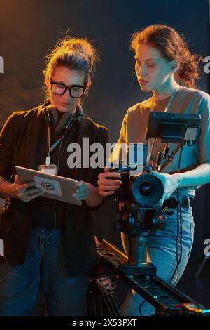 Ritratto verticale di due giovani donne che lavorano nella troupe di produzione video e guardano un tablet digitale nelle luci di studio Foto Stock