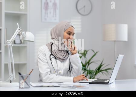 Un'allegra dottoressa musulmana in hijab si impegna con una paziente online, seduta con fiducia alla sua scrivania in una clinica moderna ben illuminata. Foto Stock