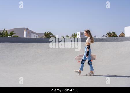 Una giovane donna principiante cammina casualmente mentre tiene in mano uno skateboard con protezioni di sicurezza Foto Stock