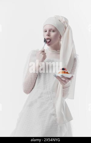 Donna che indossa un abito ricamato bianco e un elegante turbante, come una persona medievale che mangia dolci dolci sullo sfondo bianco dello studio. Foto Stock