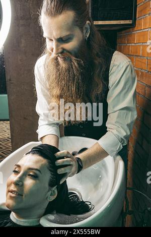 Parrucchiere che lava i capelli di una donna. Uomo giovane parrucchiere professionista esperto in gilet con barba lunga e capelli, capelli in lavaggio testa di capelli cliente femminile Foto Stock