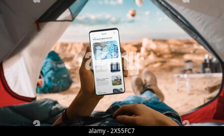 Il POV di un turista sta sta in tenda e utilizza lo smartphone per controllare le ultime notizie su Internet. Viaggiatore che riposa in tenda sulla cima di una montagna rocciosa e mongolfiere volanti. Foto Stock