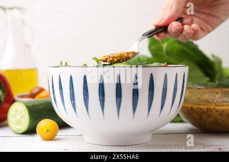 Donna che versa una gustosa salsa a base di aceto (Vinaigrette) dal cucchiaio alla ciotola con insalata al tavolo rustico in legno, primo piano Foto Stock