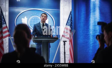 Giovane rappresentante dell'organizzazione che risponde alle domande dei giornalisti alla conferenza stampa. Addetto stampa che lavora alla riunione del Summit. Ministro che parla al Congresso. Sfondo con bandiere americane. Foto Stock