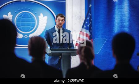 Giovane rappresentante dell'organizzazione che risponde alle domande dei giornalisti alla conferenza stampa. Addetto stampa che lavora alla riunione del Summit. Ministro che parla al Congresso. Sfondo con bandiere americane. Foto Stock