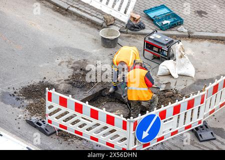 Lavori di costruzione e installazione. I lavoratori che utilizzano un martello perforatore elettrico e un generatore di benzina stanno riparando una fogna situata su una strada urbana Foto Stock