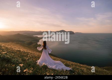 Una donna con un abito bianco si erge su una collina che domina un corpo d'acqua. La scena è serena e pacifica, con l'abito della donna che brilla nel vento Foto Stock