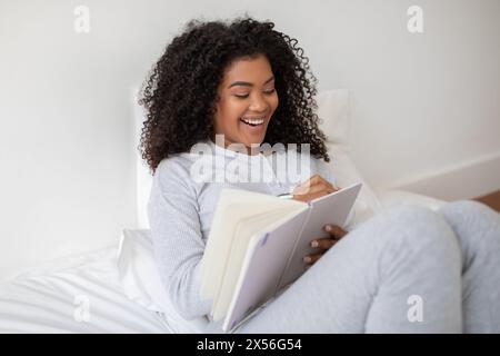 Giovane donna sorridente mentre si rilassa, scrivendo nel notebook a letto Foto Stock