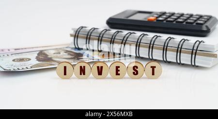 Business e finanza. Su una superficie alta si trovano un blocco note, una calcolatrice, dollari e cerchi di legno con l'iscrizione - INVEST Foto Stock